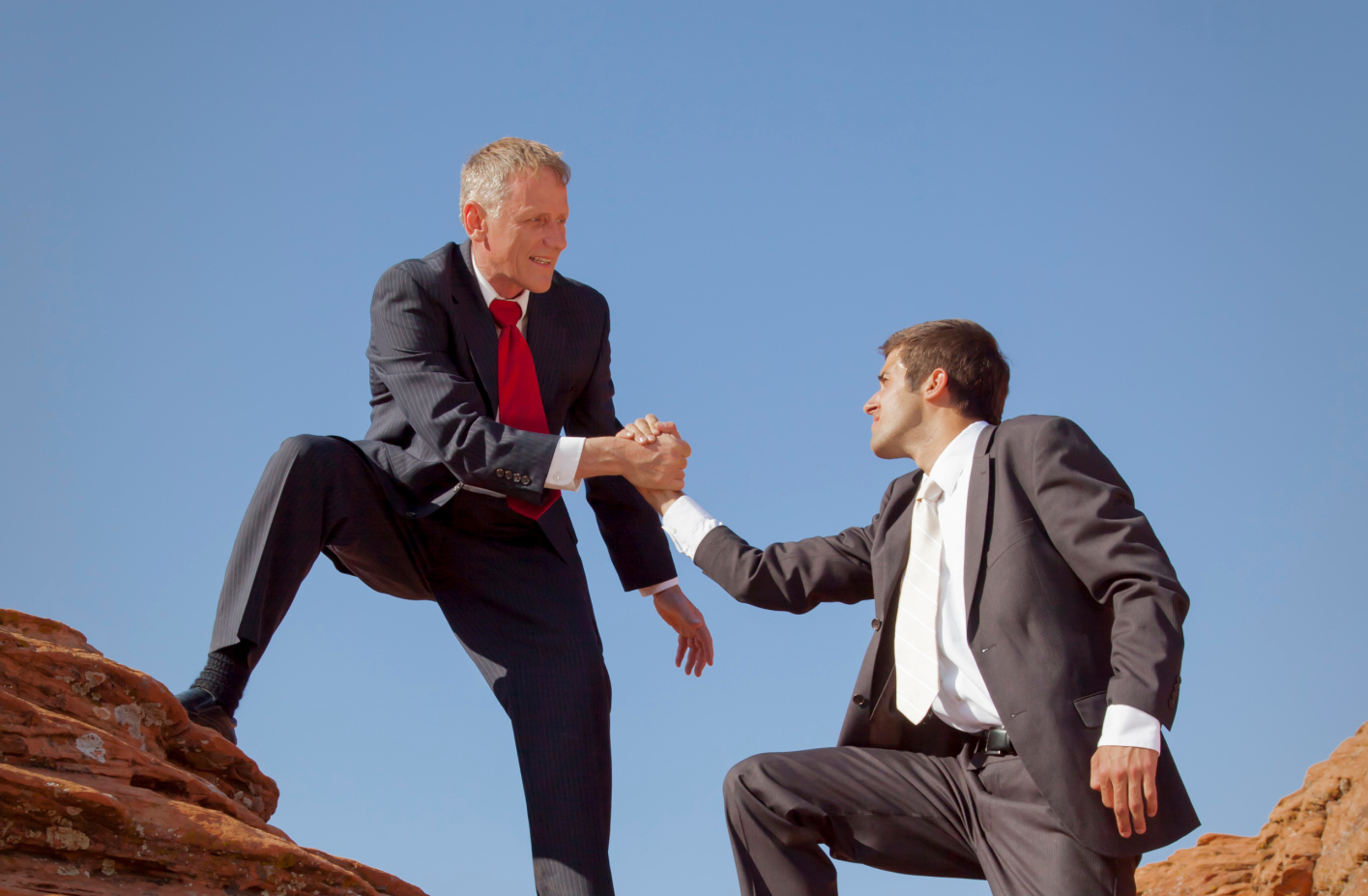  Two businessmen in suits, one on top of a rock and one below him reaching up to help him illustrates the challenges business leaders face in adopting AI.