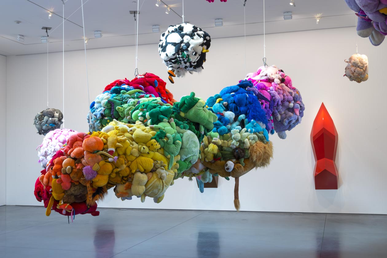 Rainbow color-coordinated stuffed animals form an amorphous glob that hangs from a ceiling.